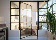 A glass and steel balcony door opens to a small balcony filled with teak chairs and coffee table atop a black bordered outdoor rug surrounded by a shiplap balcony railing.