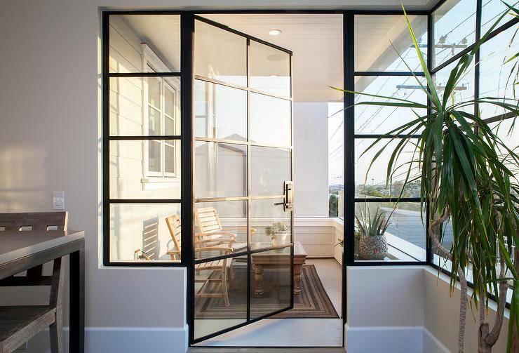 A glass and steel balcony door opens to a small balcony filled with teak chairs and coffee table atop a black bordered outdoor rug surrounded by a shiplap balcony railing.