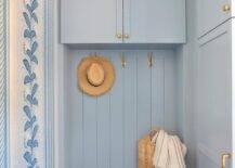 Blue mudroom features brass hooks fixed to blue shiplap trim beneath white shaker cabinets donning brass knobs and over a blue built-in storage bench finished with brass pulls. Walls are covered in