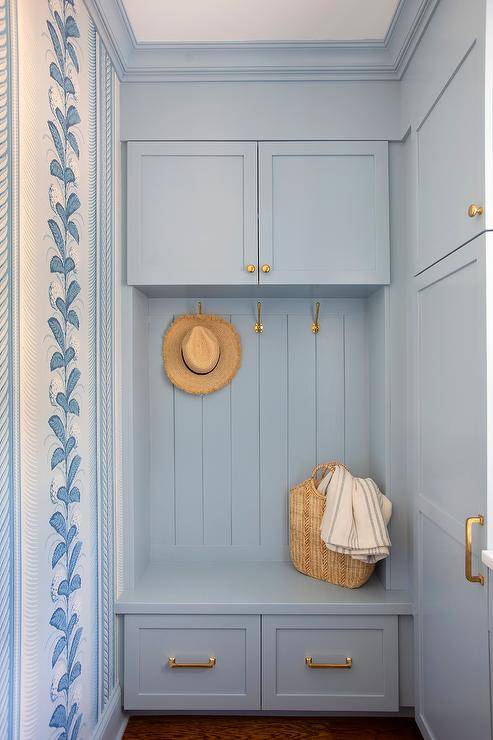 Blue mudroom features brass hooks fixed to blue shiplap trim beneath white shaker cabinets donning brass knobs and over a blue built-in storage bench finished with brass pulls. Walls are covered in