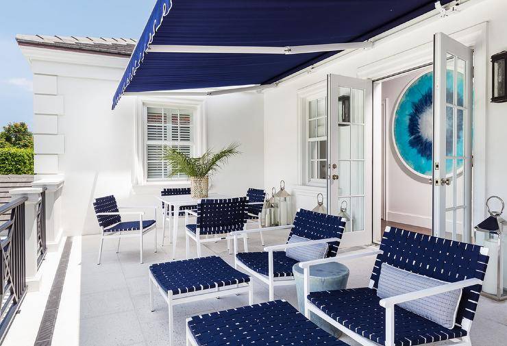 Under a navy blue balcony awning, navy blue basketweave chairs with matching stools are topped with blue stripe lumbar pillows and navy blue basketweave dining chairs surround a white dining table.
