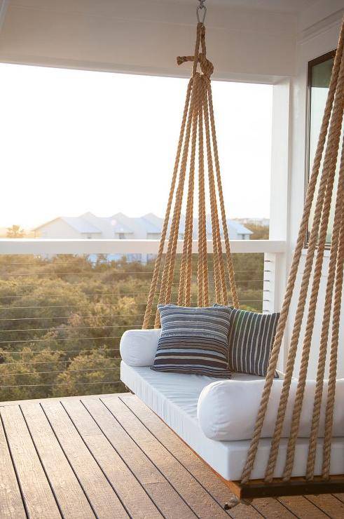 Chic covered second floor balcony is fitted with a rope swing bed adorned with plush white cushions and blue striped pillows.
