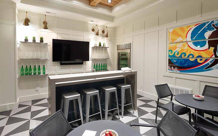 Basement wet bar design features a gray waterfall bar with tolix stools on black and white geometric floor tiles, a tv flanked by floating shelves on board and batten trim and black chairs at a black round table.