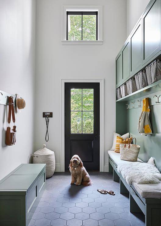 Two-story mudroom boasts black hexagon floor tiles fixed beneath a green built-in bench placed under nickel coat hooks mounted to a green plank and a green bench topped with a white cushion and whtie sheepksin throw. Fixed against a green backsplash, a row of nickel hooks are located under a green shelf holding metal bins.