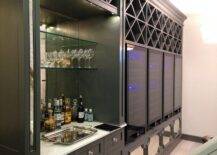 Basement bar designed with glass bar shelves and a mirrored backsplash in a dark gray built-in cabinet. A row of mirrored panels bring an elegant touch to the built-in under glass front wine cooler doors and x trim shelves.