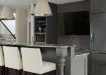 Sleek basement bar features black cabinets paired with gray granite countertops and a gray geometric tile backsplash. A flat panel tv is flanked by shelves and a microwave to the left and a dark gray paneled refrigerator to the right. Contemporary bar boasts a pair of Altamont Metal Pendants illuminating a gray bar island fitted with a sink and lined with cream leather bar stools.