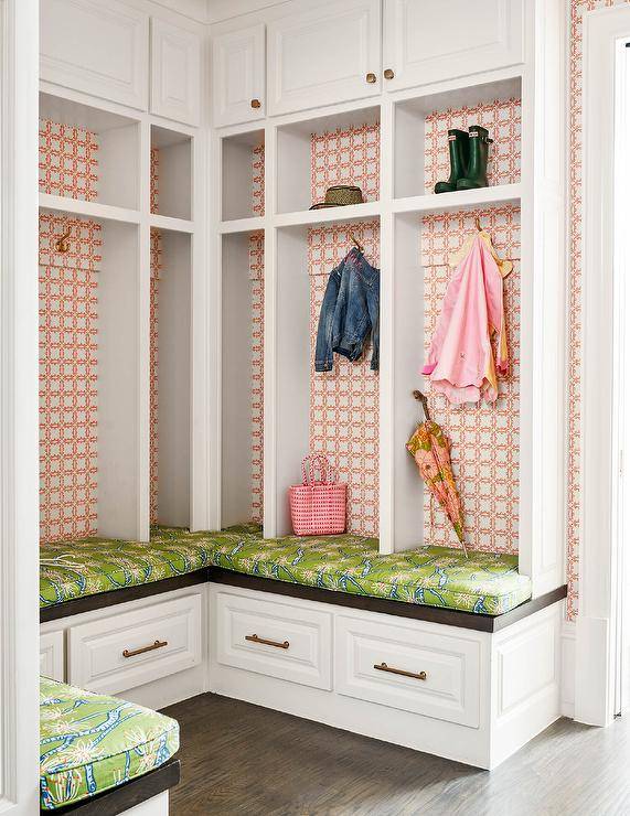 A u-shaped mudroom storage bench topped with a green and blue cushions fixed beneath white open lockers accented with pink lattice wallpapered backs. Coat hooks are mounted beneath shelves located under white overhead cabinets.