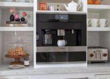 White floating shelves are stacked against white subway backsplash tiles over white cabinets topped with a honed marble countertop and on either side of a built-in Miele coffee machine. The coffee machine is fixed under white shelves and over a black oven.