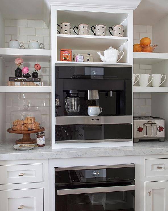 White floating shelves are stacked against white subway backsplash tiles over white cabinets topped with a honed marble countertop and on either side of a built-in Miele coffee machine. The coffee machine is fixed under white shelves and over a black oven.