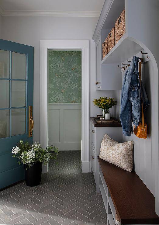 Slate herringbone patter floor tiles lead to a wooden mudroom bench fixed over gray shelves and against gray shiplap trim holding coat hooks under a gray shelf filling with woven bins. A turquoise blue mudroom door is finished with a brass door handle and glass panels.