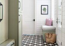 Black and white geometric floor tiles frame a green mudroom bench topped with a blue striped cushion and a white and red pillows placed against shiplap trim beneath a window. Green mudroom cabinets are stacked and adorned with oil rubbed bronze hardware.