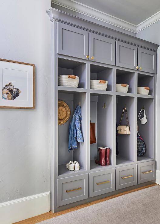 A gray mudroom boasts a row of gray open lockers along with cubbies and drawers adorned with brass pulls.