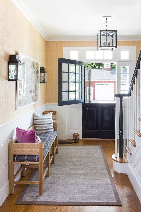 Foyer features a black dutch door with glass panels, a natural woven bench with gray cushion against gold raffia wallpaper and white tongue and groove walls over a gray bound sisal rug, lit by a black framed lantern.