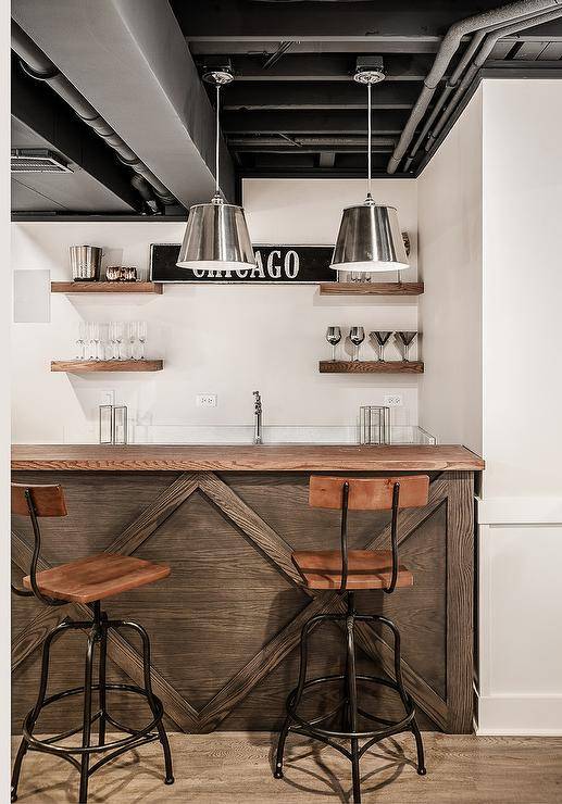 An industrial style basement features a distressed coffee stained oak wood bar counter with metal and wood swivel stools under silver metal light pendants. Stained oak floating wet bar shelves flank 