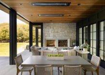 Covered patio with ceiling heaters on ceiling planks features an gray outdoor dining table with gray rope outdoor dining chairs and a sitting area with fireplace.