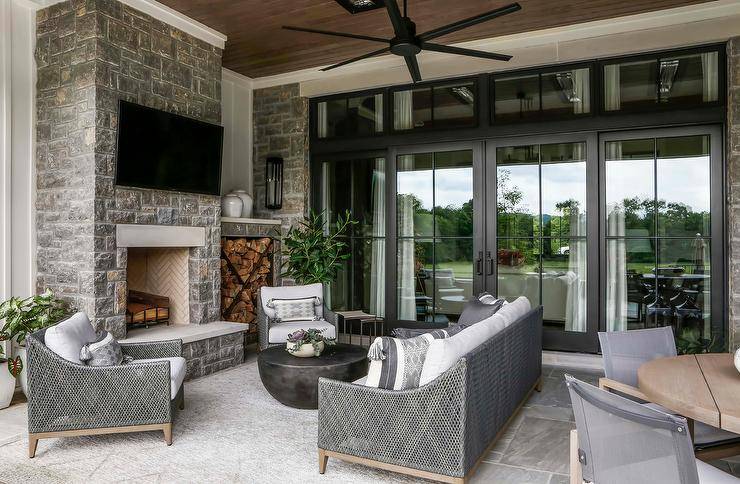 A gray wicker sofa accented with gray pillows sits on a wonderfully appointed covered patio facing a round black coffee table flanked by gray wicker lounge chairs. A flat panel tv is mounted to a gray stone fireplace located next to custom fire wood cubbies.