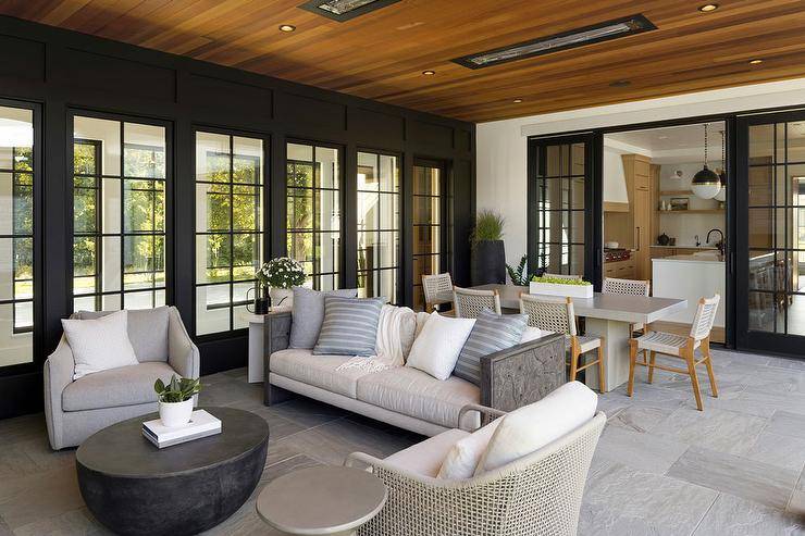 Covered patio features a gray outdoor swivel chair with round black outdoor coffee table, a gray and white outdoor sofa, a gray woven outdoor chair and ceiling heaters on ceiling planks over an gray outdoor dining table with gray rope outdoor dining chairs.
