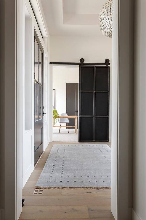 A beautifully styled entryway, a gray rug sits atop a gray wash wide plank wood floor and leads to a home office boasting an industrial metal door on rails.