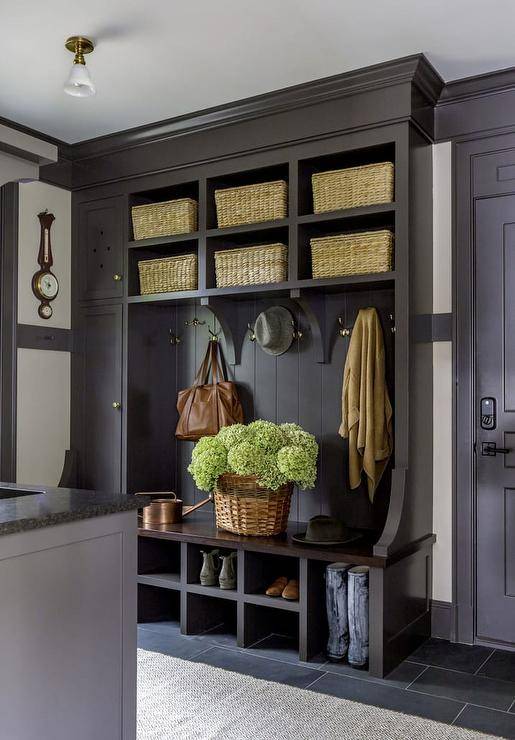 Black cottage style mudroom features a black built-in bench fitted iwth shoe shelves and mounted against black shiplap trim holding polished nickel hooks under stacked black shelves holding woven bins. A gray runner covers black slate floor tiles.