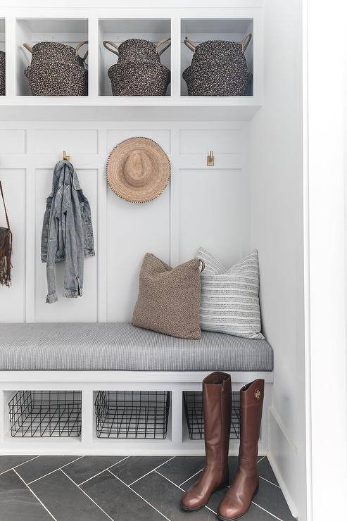 Gray herringbone tiles lead to a built-in mudroom bench fitted with cubbies holding metal bins and positioned beneath a gray cushion. The bench is mounted against a white board and batten trim holding brass hooks beneath overhead cubbies stuffed with woven baskets.