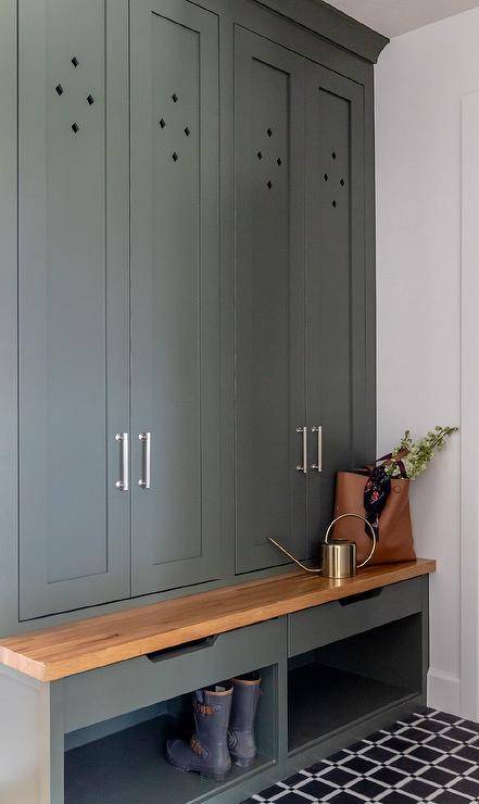 Gray-green shaker mudroom cabinets are accented with polished nickel pulls and fixed over a wood top bench finished with drawers and shoe cubbies. The space is complemented with black and white grid floor tiles.