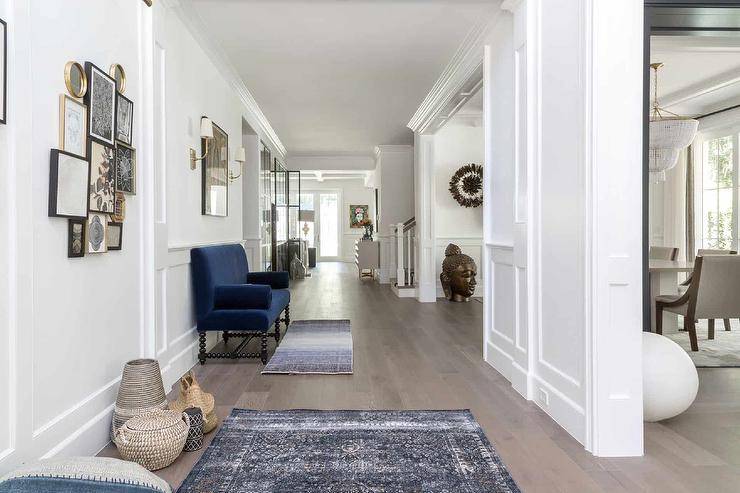 Long entry features a royal blue velvet settee with brass picture lights over an eclectic art gallery, dark blue floor runners and woven baskets.