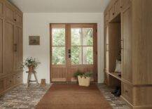 Wood and glass double doors open to reveal a cottage mudroom featuring a brown jute rug placed on red brick pavers between floor-to-ceiling light brown closed lockers and a light brown wooden stool framed by light brown cabinets with vintage bronze hardware.