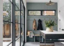 Black sliding glass doors open to a beautifully designed mudroom featuring an industrial freestanding island placed against dark gray picket floor tiles perpendicular to a built-in wood top bench. The bench is fixed against gray shiplap trim holding brass hooks beneath windows.