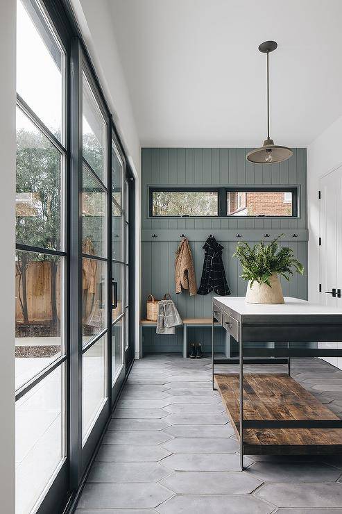 Black sliding glass doors open to a beautifully designed mudroom featuring an industrial freestanding island placed against dark gray picket floor tiles perpendicular to a built-in wood top bench. The bench is fixed against gray shiplap trim holding brass hooks beneath windows.