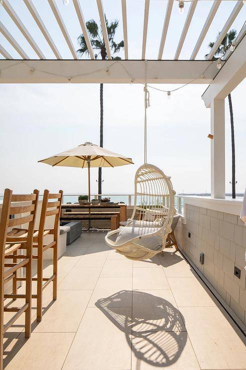 A beach cottage patio features a hanging white rattan chair hung from a white pergola.