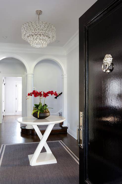 Crystal cascading chandelier illuminates a foyer furnished with a small round white trestle accent table on a dark gray rug with a white border. A glossy black front door with a silver lion door knocker brings a classic appeal to the entryway of the home.