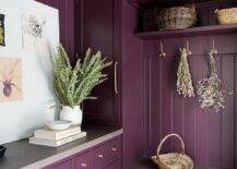 Purple mudroom design features an aubergine and purple stone bench under aubergine shiplap trim, aubergine mudroom cabinets accented with brass hardware and red brick herringbone pavers.