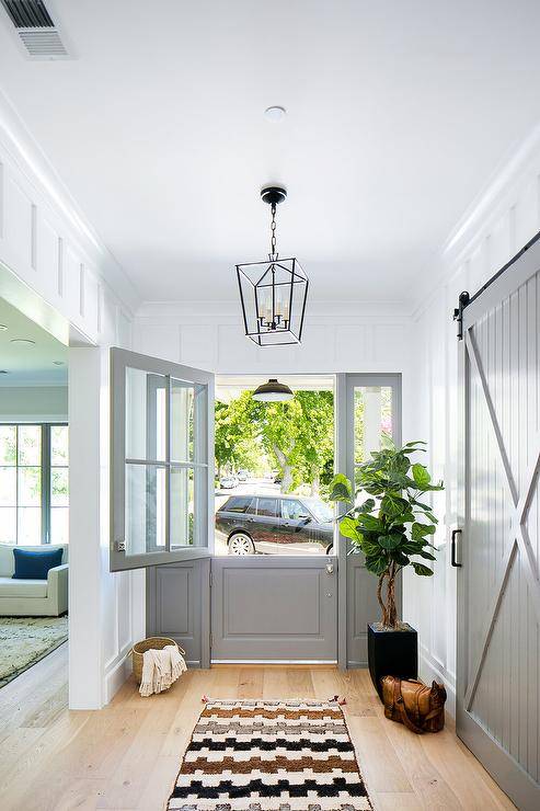 Darlana Lantern illuminates a front entryway designed with a gray dutch front door and glass side panels. Light oak wood floors are styled with baskets, decor and a vintage rug in black, brown and cream.