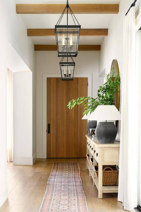 Entry features a cream console table lit by black urn lamps, a pink and gray vintage runner illuminated by bronze lanterns on foyer ceiling beams and a plank front door.