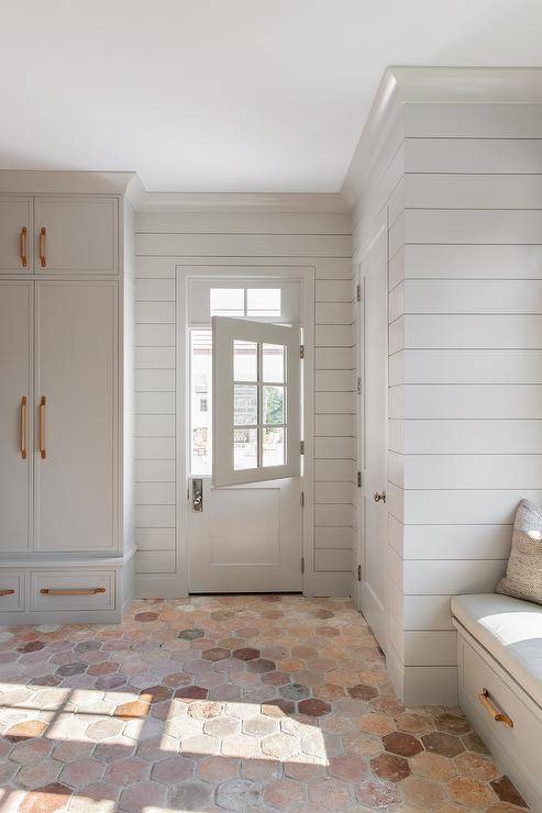 Light gray closed mudroom lockers are fixed against terracotta hexagon pavers beside a light gray Dutch door framed by light gray shiplap. A light gray built-in bench is topped with a light gray cushion and finished with wood and leather pulls.