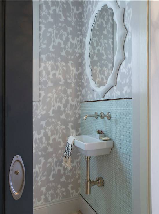 Chic powder room features a black door opening to reveal subtle white and gray floral motif wallpaper highlighting a modern white mirror mounted above an aqua blue glass mini brick tiled backsplash trimmed with black pencil tile framing a mini wall mount sink with modern wall mount faucet.