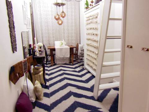 A crash pad with a white ladder leading up to a Pied-A-Terre, featuring blue chevron carpet tiles, a small wood table, chairs, and throw pillows in varying colors on the floor.