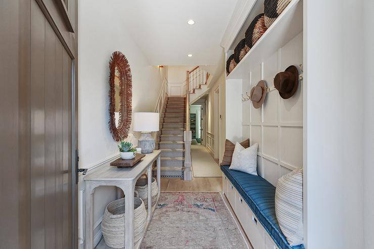 Charming narrow foyer features a long white built-in bench with a blue cushion, a brown leather oval mirror over a gray wash console table with gray woven baskets and a red and gray faded rug.