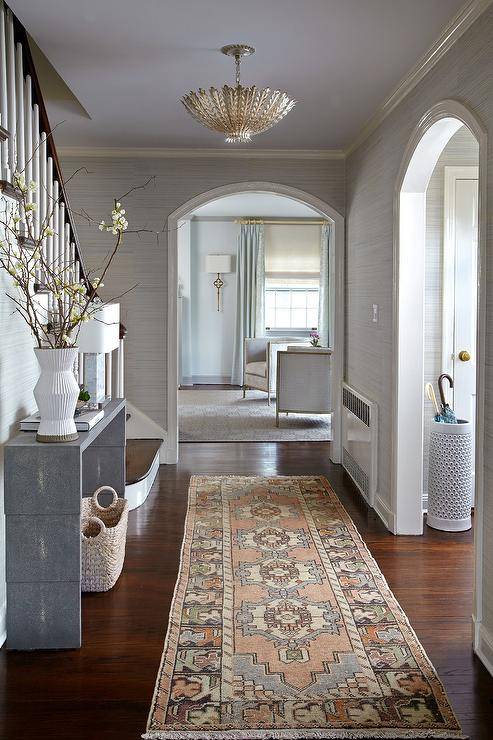 Foyer features a pink and blue vintage runner in front of a gray waterfall console table against a stairwell lit by a chandelier.