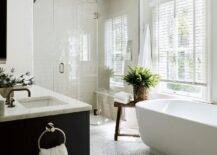 An oval freestanding bathtub is placed on recycled floor tiles beside a rustic wooden stool positioned beneath a window covered in white blinds and beside a frameless glass shower.