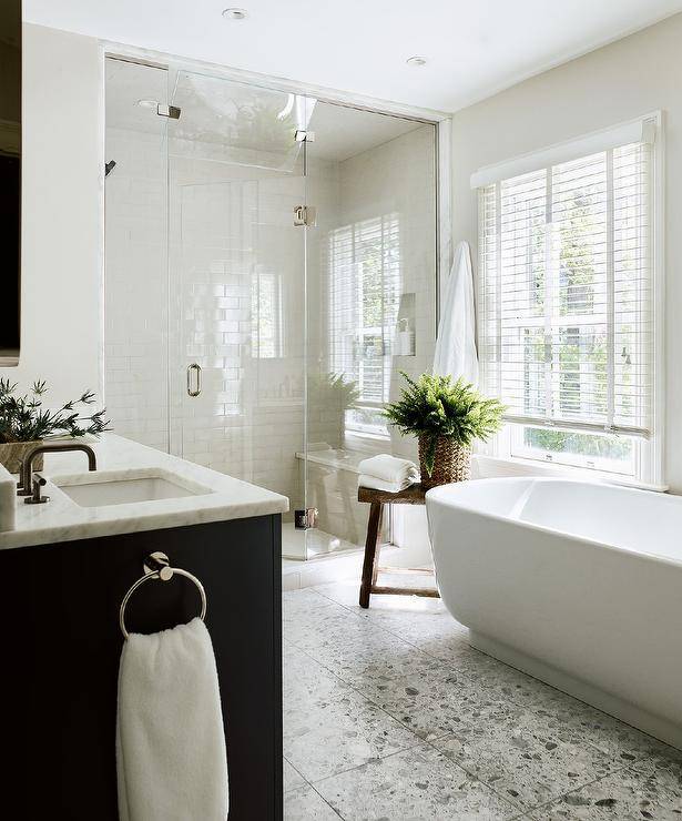 An oval freestanding bathtub is placed on recycled floor tiles beside a rustic wooden stool positioned beneath a window covered in white blinds and beside a frameless glass shower.