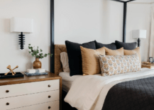 A black wooden canopy bed is fitted with a brown abaca headboard and dressed in black and brown bedding. An oil-rubbed bronze sconce sits over an ivory and brown dresser adorned with brass knobs.