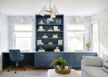 Windows dressed in white roman shades framed by gray and blue wallpaper flank a blue bookcase fixed atop a long blue desk. The desk is finished with blue cabinets donning brass hardware and paired with a blue leather task chair placed on a blue rug.