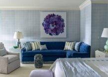 Bedroom features a sitting area with a blue velvet tufted sectional under purple floral art, a blue chinoiserie end table lit by a blue lamp and a gray gingham chair.