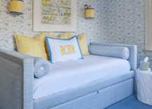 Nickel sconces with yellow and blue shades flank an art piece hung from a blue wallpapered wall over a blue daybed accented with blue pillows. The bed sits in a yellow and blue nursery on a blue rug.