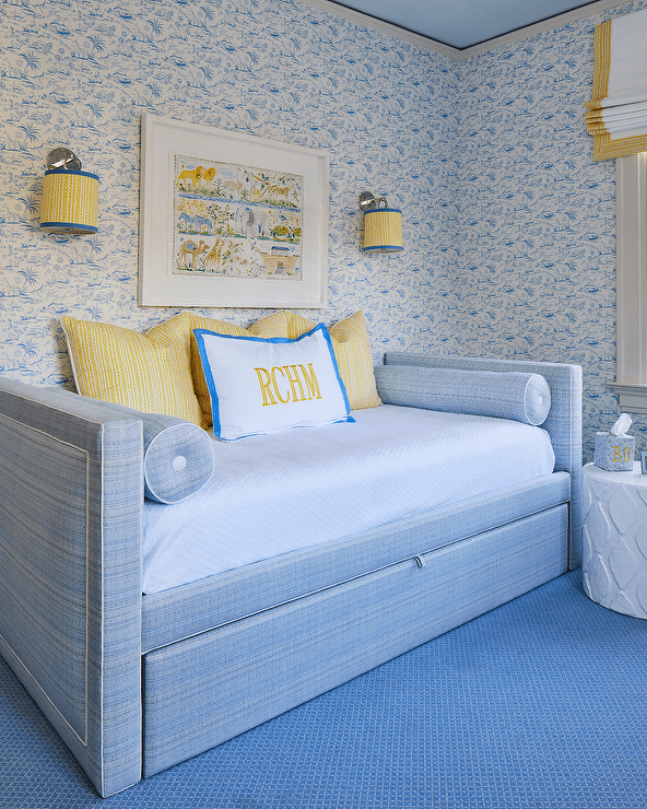 Nickel sconces with yellow and blue shades flank an art piece hung from a blue wallpapered wall over a blue daybed accented with blue pillows. The bed sits in a yellow and blue nursery on a blue rug.