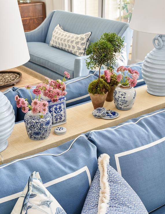 A tan raffia sofa table topped with light blue lamps and styled with white and blue chinoiserie vases sits behind a blue sofa.
