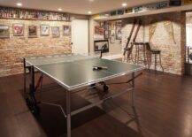 Basement man cave features a ping pong table placed in the center of a room clad in exposed brick and fitted with a long floating shelf showcasing stacked bear cans and fixed above framed photographs. A tall round pub tables seats two backless stools against a wall accented with antique sports gear and beneath a row of license plates.