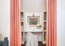 Salmon pink curtains cover a kid's desk nook featuring a skirted desk chair placed at a white built-in desk fixed beneath a lighted art piece hung from a wall clad in shiplap trim. The desk is flanked by white shelves.