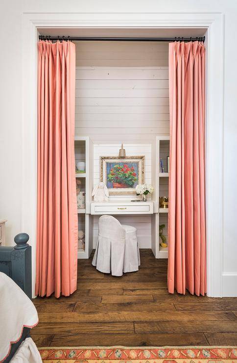 Salmon pink curtains cover a kid's desk nook featuring a skirted desk chair placed at a white built-in desk fixed beneath a lighted art piece hung from a wall clad in shiplap trim. The desk is flanked by white shelves.